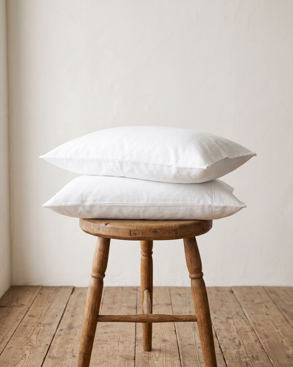 Two white queen and king pillows cases stacked on a rustic wooden stool in a minimalist room with a cream wall and wooden floor.