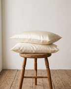Two ivory queen and king pillows stacked on a rustic wooden stool in a room with a white wall and wooden floor.